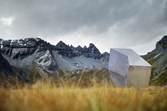 Eine Metallskulptur steht auf einer Wiese mit Bergen im Hintergrund.