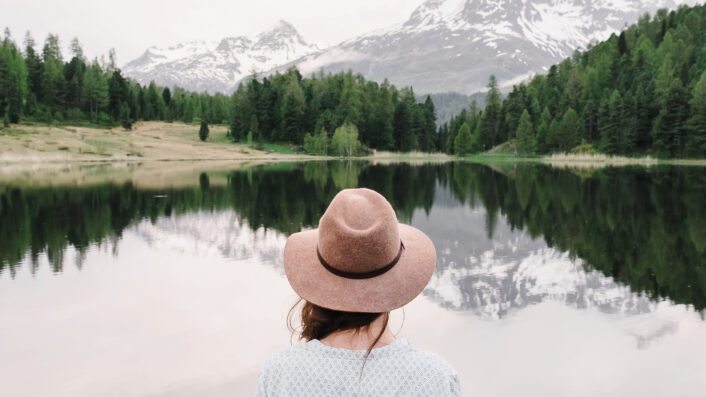 Eine Frau mit Hut blickt auf einen See mit Bergen im Hintergrund.